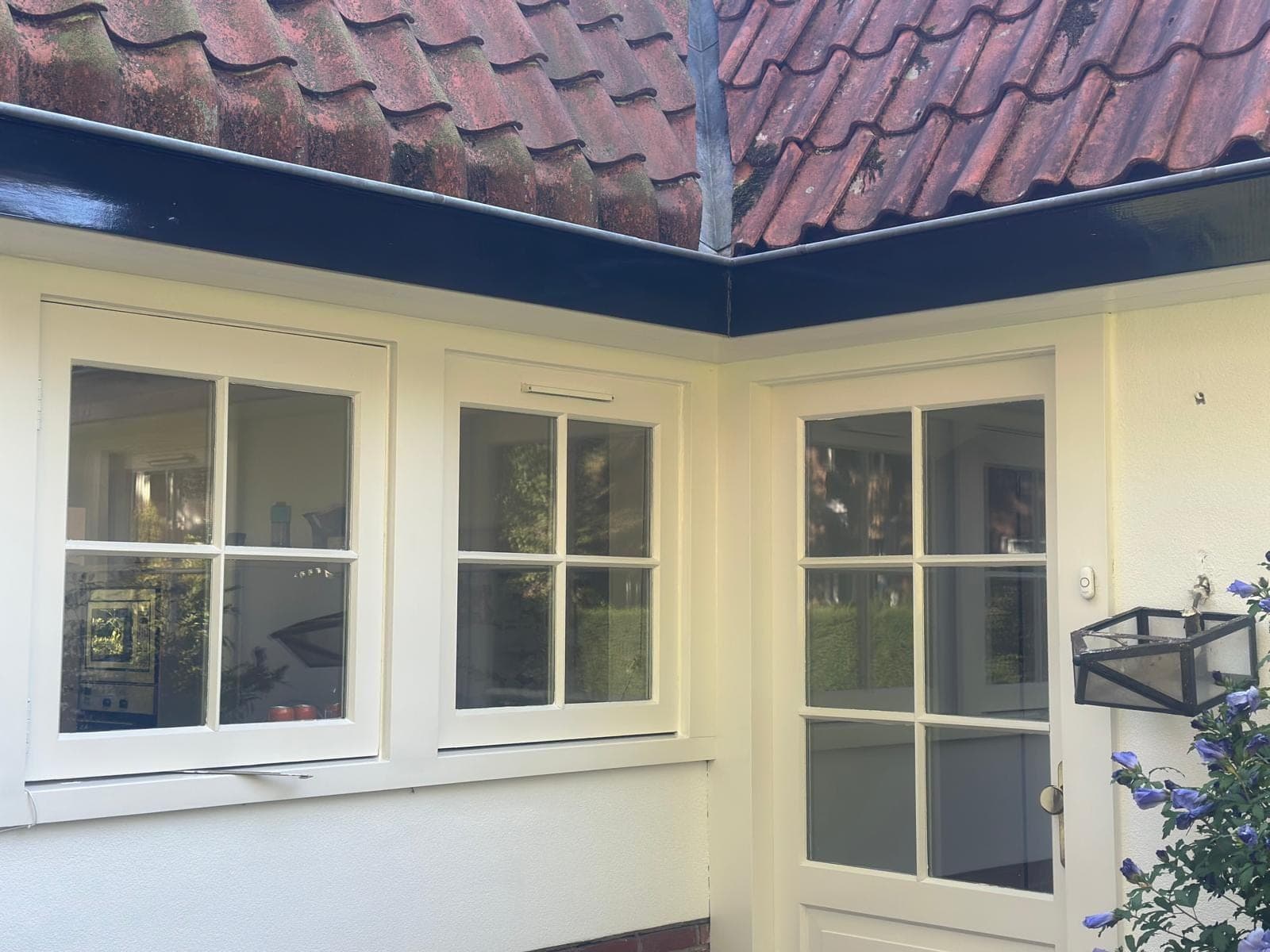 Hoek van de woning met crèmewitte kozijnen, een deur met roedes en een diepblauwe dakgoot onder rode pannen