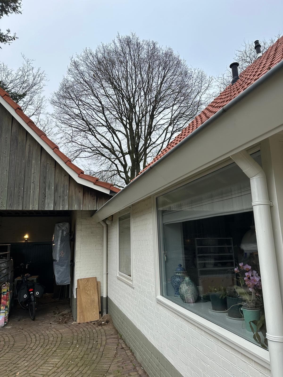 Zijgevel van de boerderijwoning met een groot wit kozijn, witte regenpijp en de open schuur